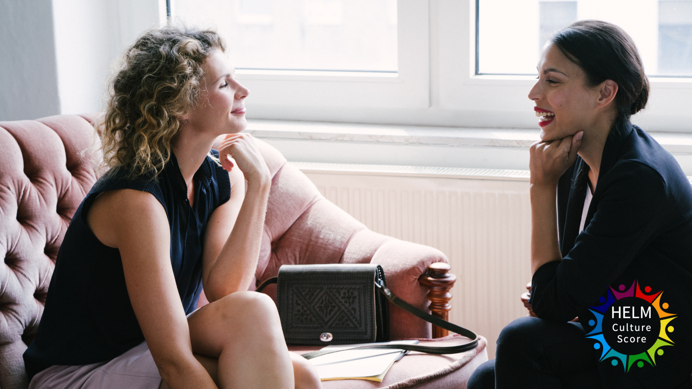 Two businesswomen in conversation on a sofa illustrating mentoring vs coaching, with HELM Culture Score logo